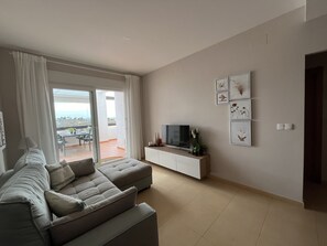 Un salon moderne avec un canapé d’angle, un téléviseur à écran plat posé sur une console blanche et un balcon offrant une vue agréable.