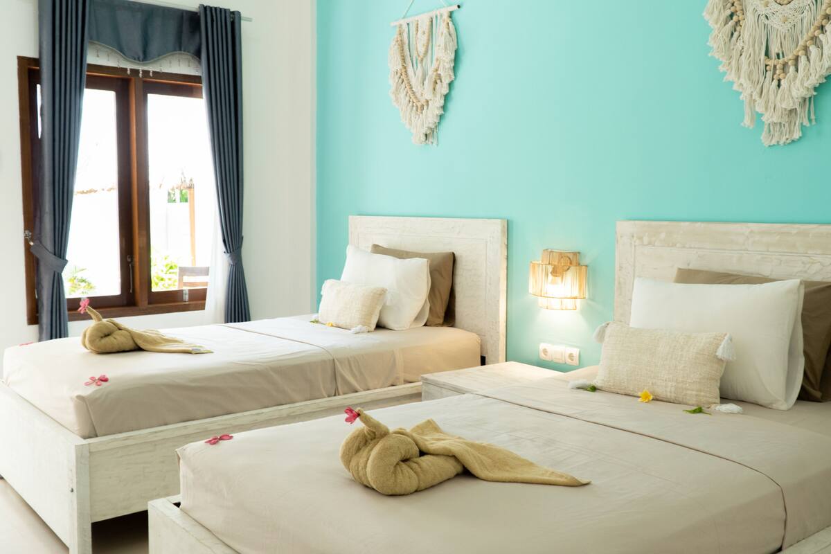 Habitación de hotel con dos camas, una ventana con cortinas y decoración de macramé en la pared.