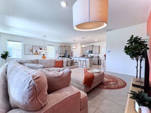 A modern living room with a large L-shaped sectional sofa, a kitchen in the background, and a dining area with bar stools.
