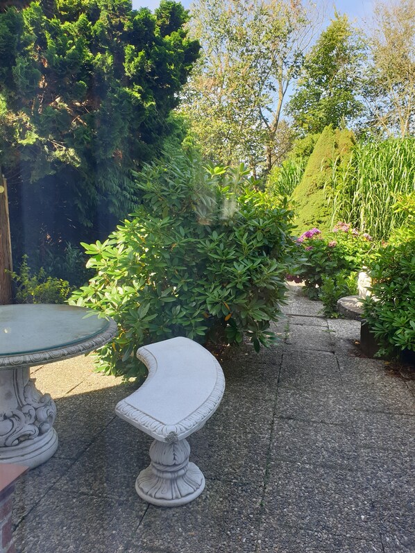 A garden pathway with a white stone bench and a table, surrounded by lush greenery and a neatly trimmed hedge.