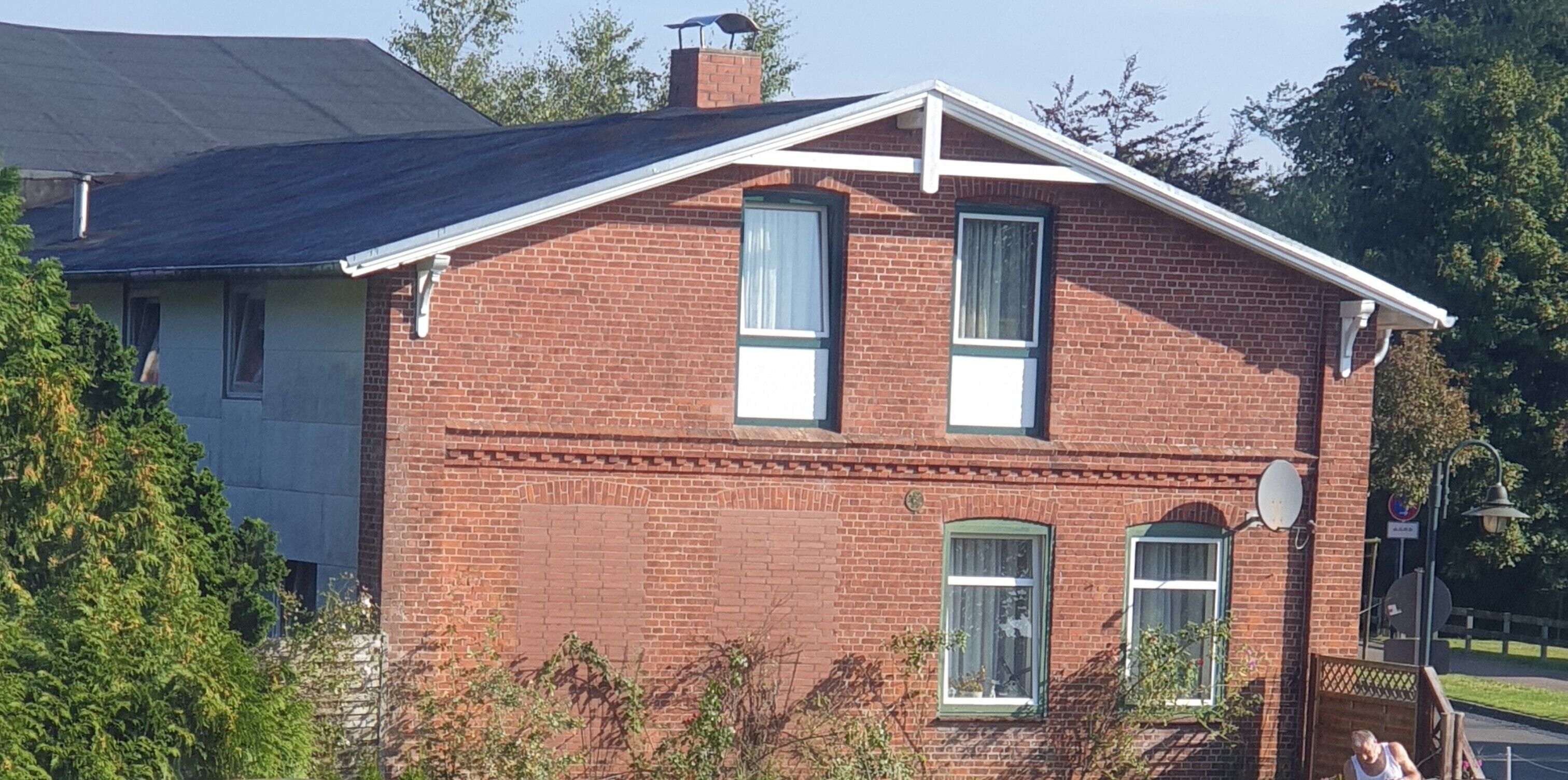 Une maison à deux étages, construite en briques, avec un toit à pignon, deux fenêtres dotées de volets et une antenne parabolique sur le toit.