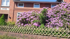 A brick building with a number '1918' on the facade, a wooden fence, and purple flowering bushes. - Ferienwohnung in Alleinnutzung mit Garten und Teich, Nähe Sankt Peter Ording (Lehe)