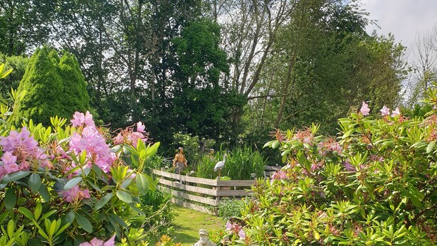 A garden with a statue of a man holding a basket, surrounded by lush greenery and pink flowers. - Ferienwohnung in Alleinnutzung mit Garten und Teich, Nähe Sankt Peter Ording (Lehe)