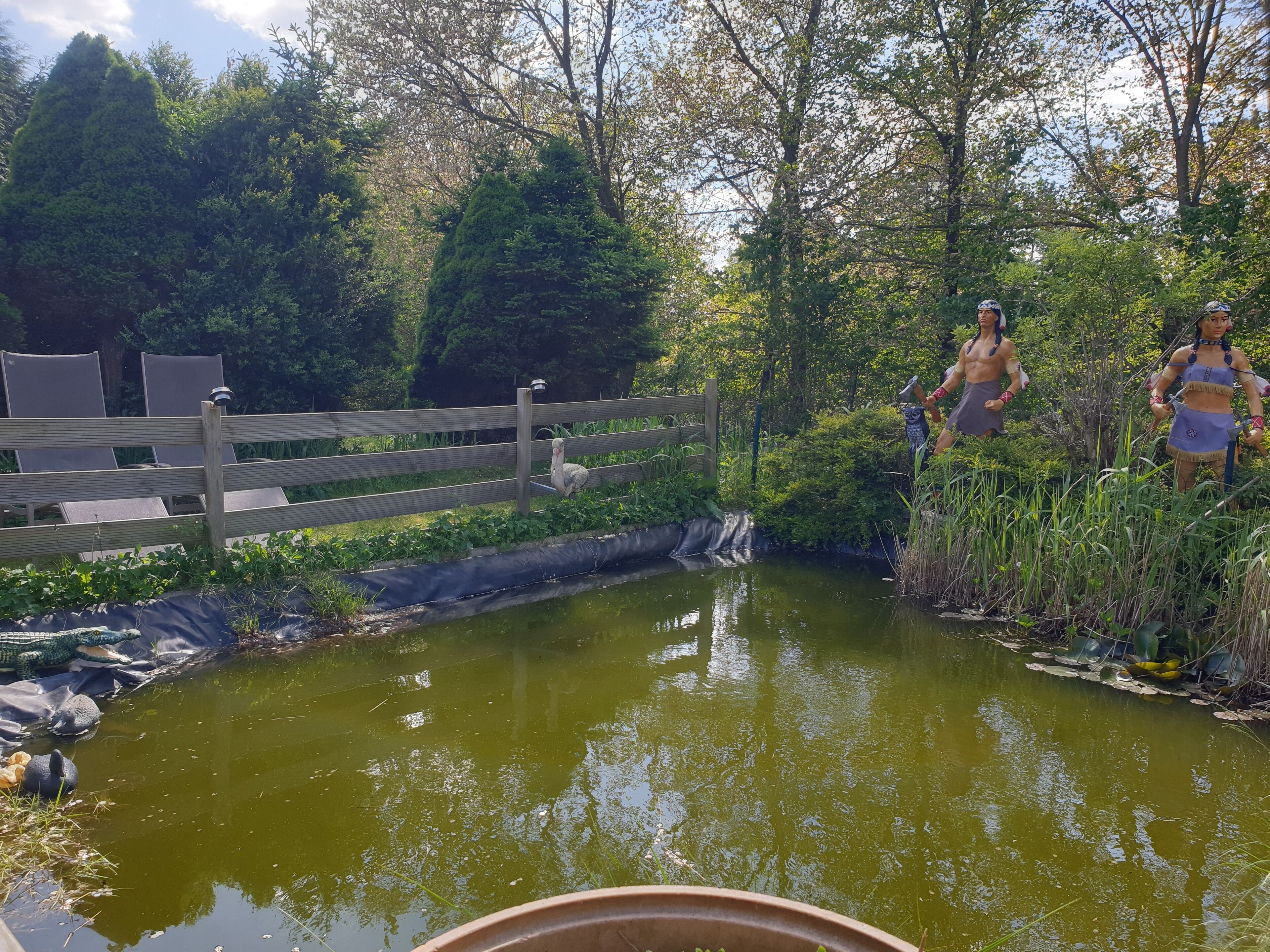 Un puente de madera sobre un estanque, rodeado de follaje y con una estatua.