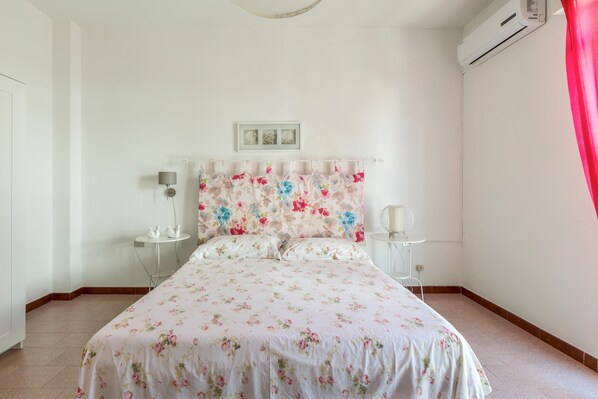 Une chambre avec une couvre-lit à motifs floraux, une tête de lit blanche, une table de chevet avec une lampe et un appareil de climatisation fixé au mur.