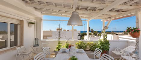 A covered outdoor dining area with a table set for two, surrounded by white chairs, under a white ceiling with exposed beams.
