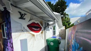 A narrow alleyway with a white building on the left, a green trash bin, and a mural on the right wall.
