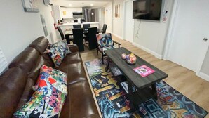 A living room with a brown leather sofa, a coffee table with colorful artwork, and a dining area with a kitchen in the background. - Unique Miami Art Escape w/ HotTub, Arcade & Murals (Miami)