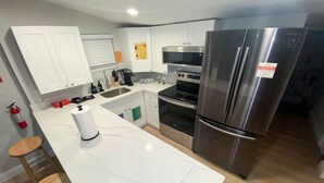 A modern kitchen with white cabinets, a stainless steel refrigerator, an oven, and a microwave. There are two wooden stools and a fire extinguisher on the wall. - Unique Miami Art Escape w/ HotTub, Arcade & Murals (Miami)