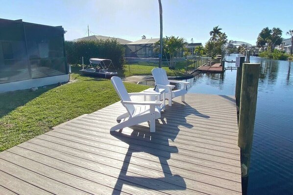 A wooden deck with white chairs and a table, overlooking a body of water and a residential area. - Gulf Gateway-Pool/Dock/Grill/Fun (Fort Myers)