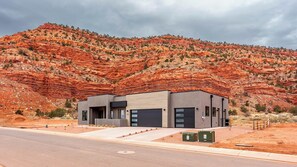 Il s’agit d’un bâtiment moderne à deux étages, doté d’un toit plat, de grandes fenêtres et d’une porte de garage. Il est situé dans un paysage désertique, avec des formations rocheuses rouges en arrière-plan.