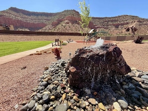 Una roca de la cual brota agua, rodeada de piedras y un área verde con bancos y un parque infantil al fondo.