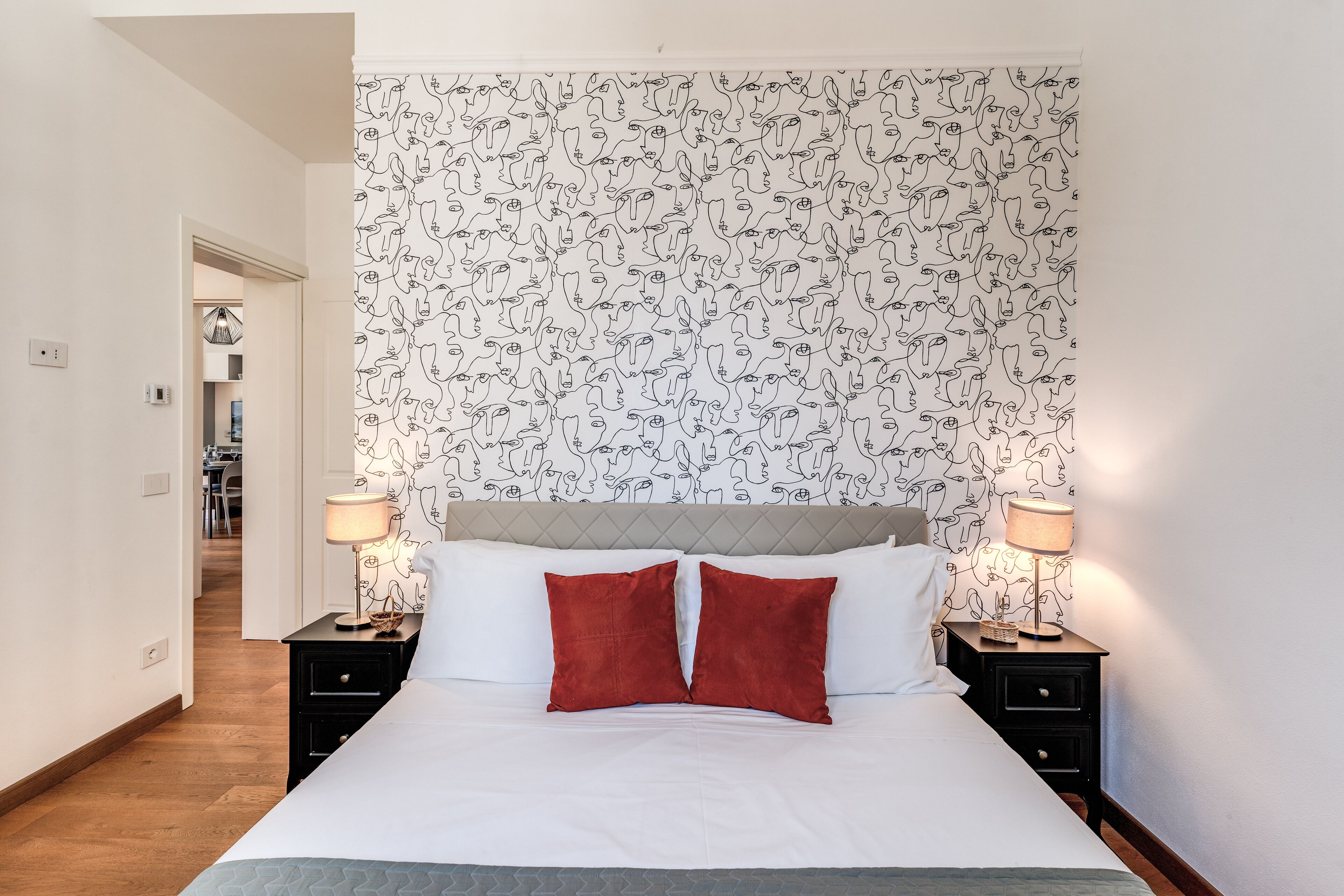 Una habitación de hotel con una cama, dos mesitas de noche, un cabecero con papel tapiz estampado y un pasillo que lleva a otra habitación.