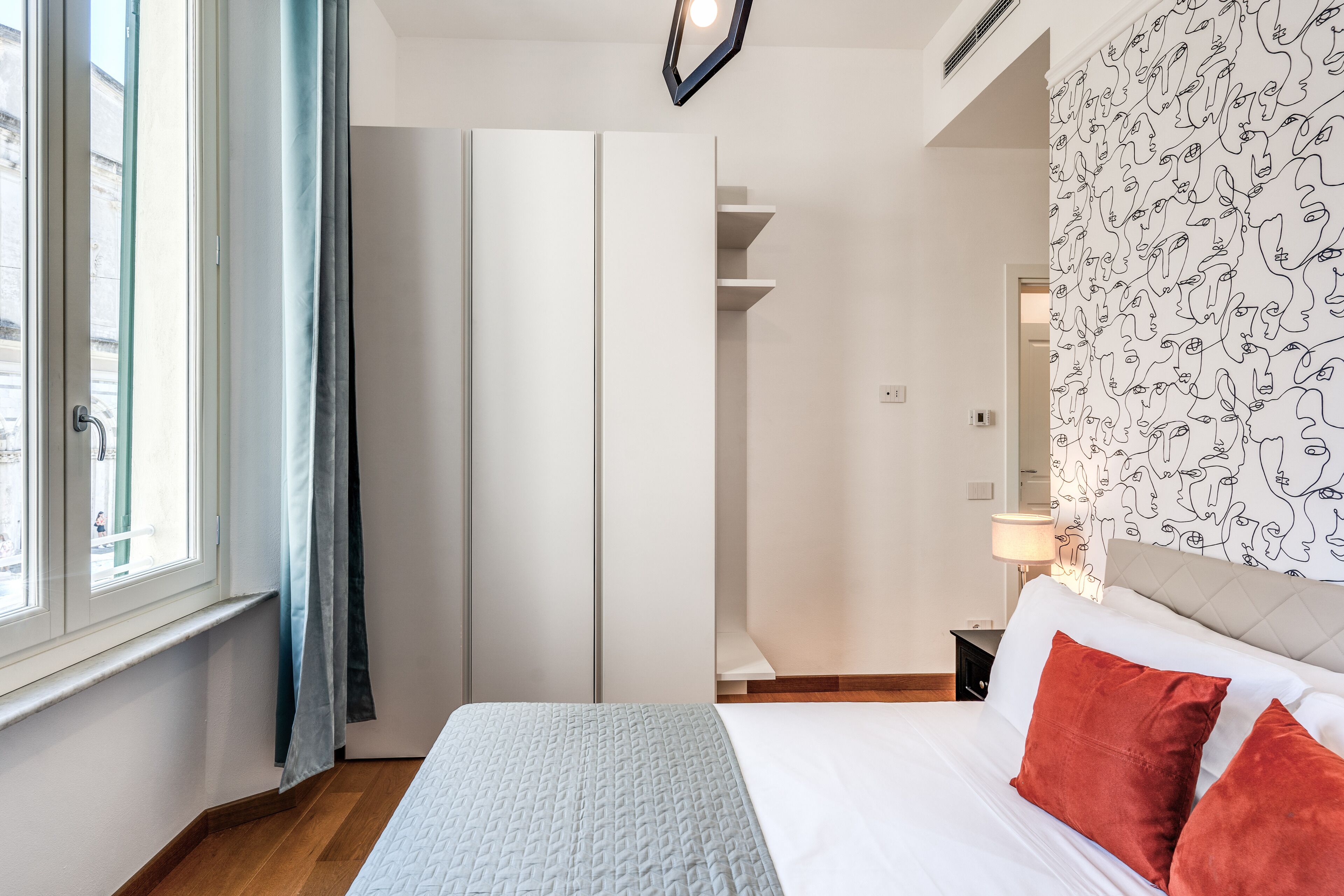 A bedroom with a bed, a nightstand, a window with curtains, and a wall with a pattern.