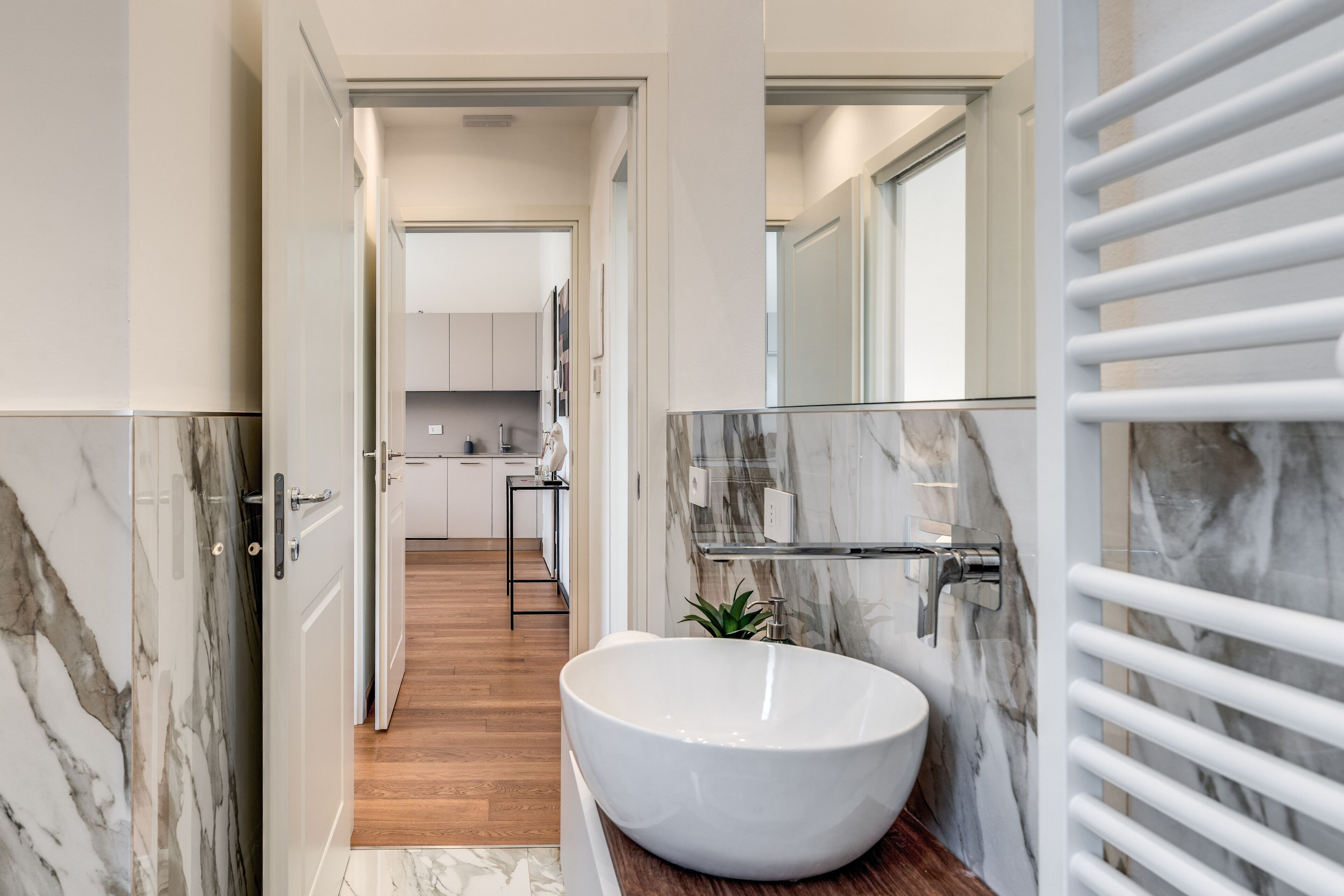 A modern bathroom with a large white vessel sink, marble walls, and a wooden floor.