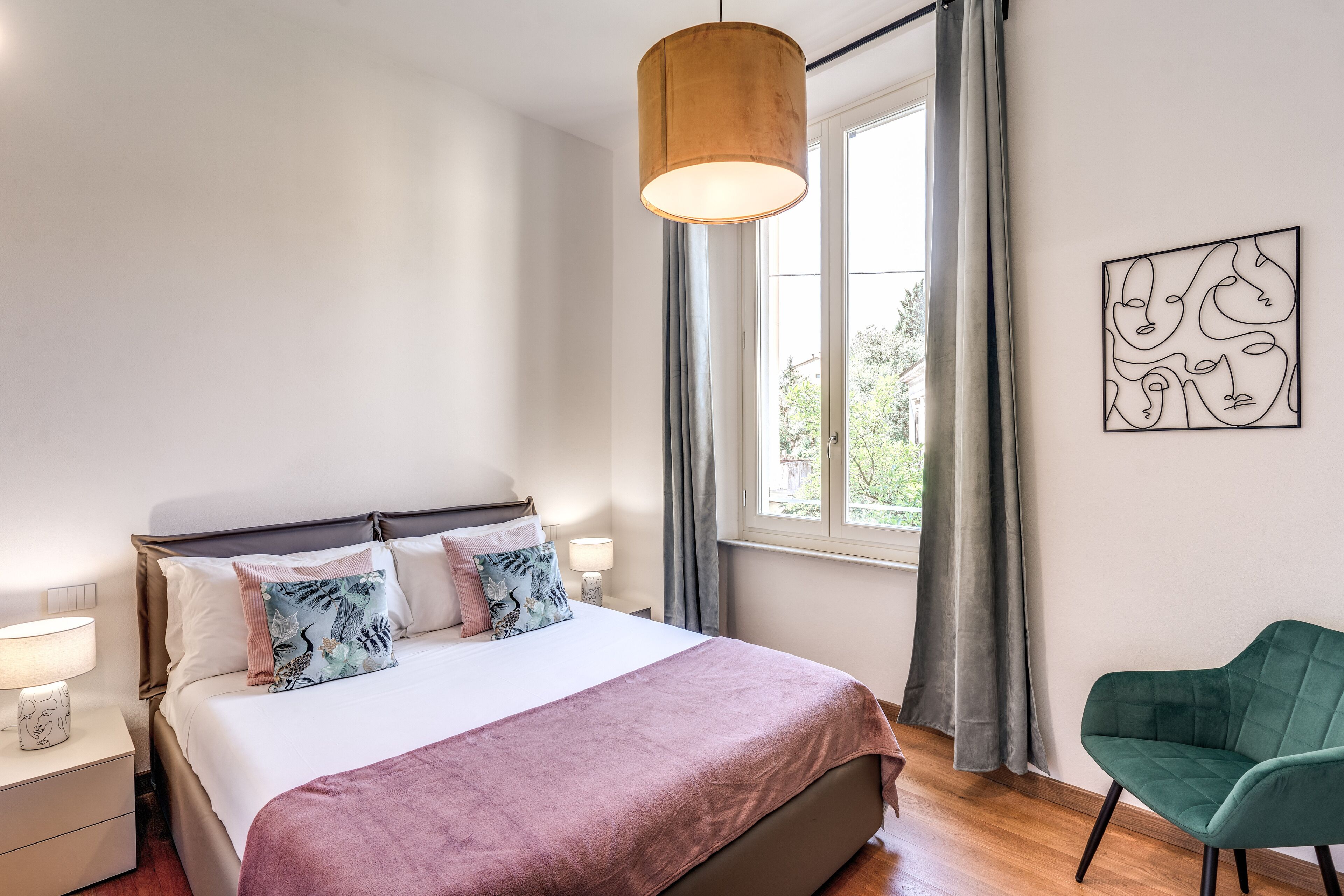 A bedroom with a bed, bedside lamps, a window with curtains, a green chair, and a wall art piece.