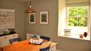 A dining area with a wooden table, chairs, a radiator, and a window with a view of trees.