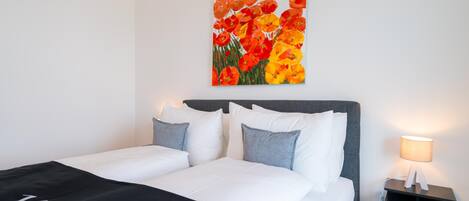A neatly made bed with white and grey bedding, a nightstand with a lamp, and a painting of flowers on the wall.