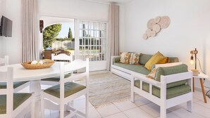 A bright, modern living room with a white sofa, green cushions, a white dining table with chairs, and a large window offering a view of the outdoors. - Flat Buganvilia OCV - Pool & Private Parking (Albufeira)