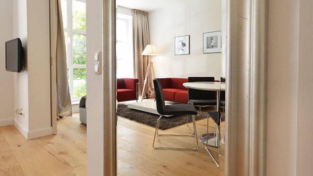 A modern hotel room with a large mirror, wooden flooring, a red sofa, a dining table, and a TV mounted on the wall. - Vienna Residence, Frankfurt - Schweizer Platz #6070 (Frankfurt)