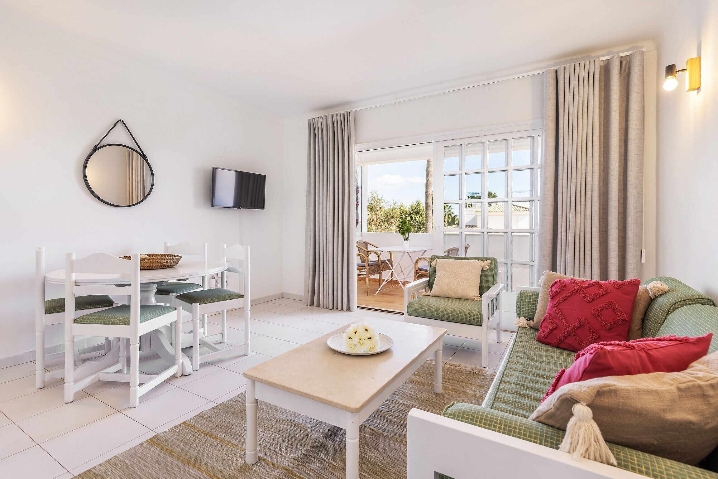 A modern living room with a dining area, a flat-screen TV, and a view through a large window.