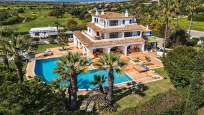 Une grande maison à deux étages, dotée d’une piscine, entourée d’une végétation luxuriante et de palmiers.
