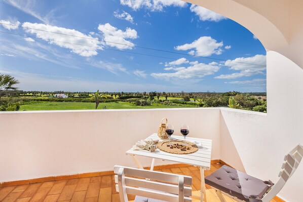 A balcony with a table set for two, a cushioned chair, and a view of a green landscape.