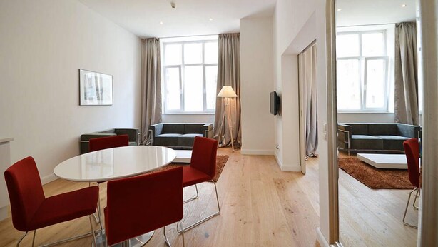 Een moderne woonkamer met een witte eettafel, rode stoelen, een grijze bank en een grote spiegel die de kamer weerspiegelt.
