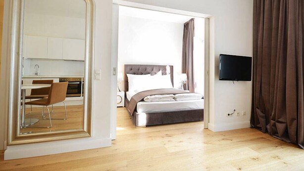 Une chambre avec un lit, une télévision, un coin repas avec une chaise, et une cuisine visible à travers une porte ouverte.