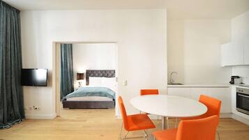 A modern hotel room with a bed, a dining table with orange chairs, a kitchen area, and a TV mounted on the wall.