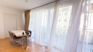 Quarto com mesa de jantar, cadeiras, vaso de flores, cortinas translúcidas e vista pela janela.