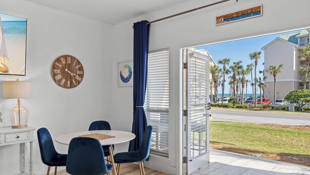 A room with a round table, blue chairs, a wooden clock, a painting, and a view of palm trees and a road outside. - Great Location with Easy Beach Access! (Destin)