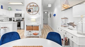 A modern kitchen with white cabinetry, a microwave, and a coffee machine. There is a dining area with a white table and blue chairs. - Great Location with Easy Beach Access! (Destin)