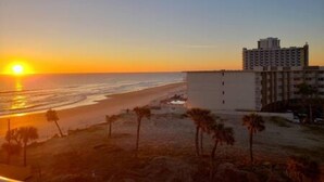 Vista de la playa con un atardecer, un edificio de varios pisos y palmeras.