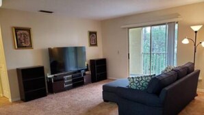 A living room with a dark blue sofa, a flat-screen TV on a stand, a large window with blinds, and a floor lamp. - 101 Bent Tree Modern Lux 2BR 2BA Heated Pool (Daytona Beach)