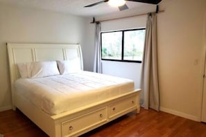 A bedroom with a bed, a ceiling fan, a window with curtains, and a wooden floor.