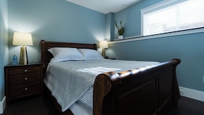 A bedroom with a wooden bed, two bedside tables, a nightstand, a window with blinds, and a potted plant on the windowsill.