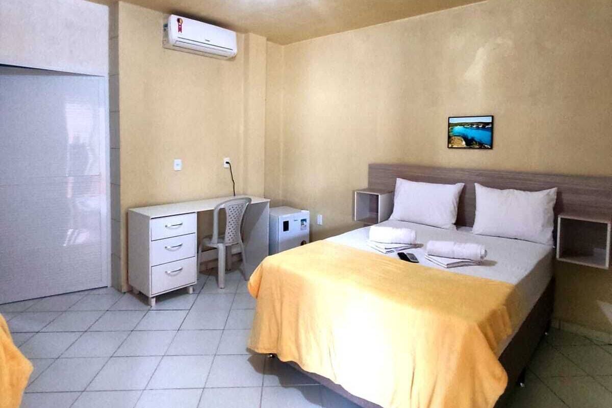 A hotel room with a bed, a desk, a chair, an air conditioning unit, and a small picture on the wall.
