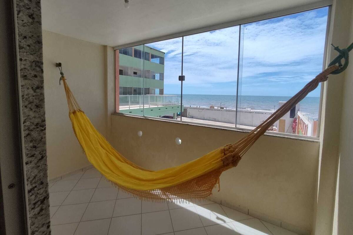 A room with a hammock, a large window, and a view of the beach and buildings.