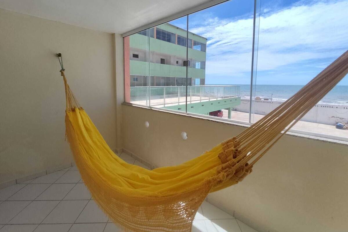A room with a hammock by a large window overlooking a beach and a building.