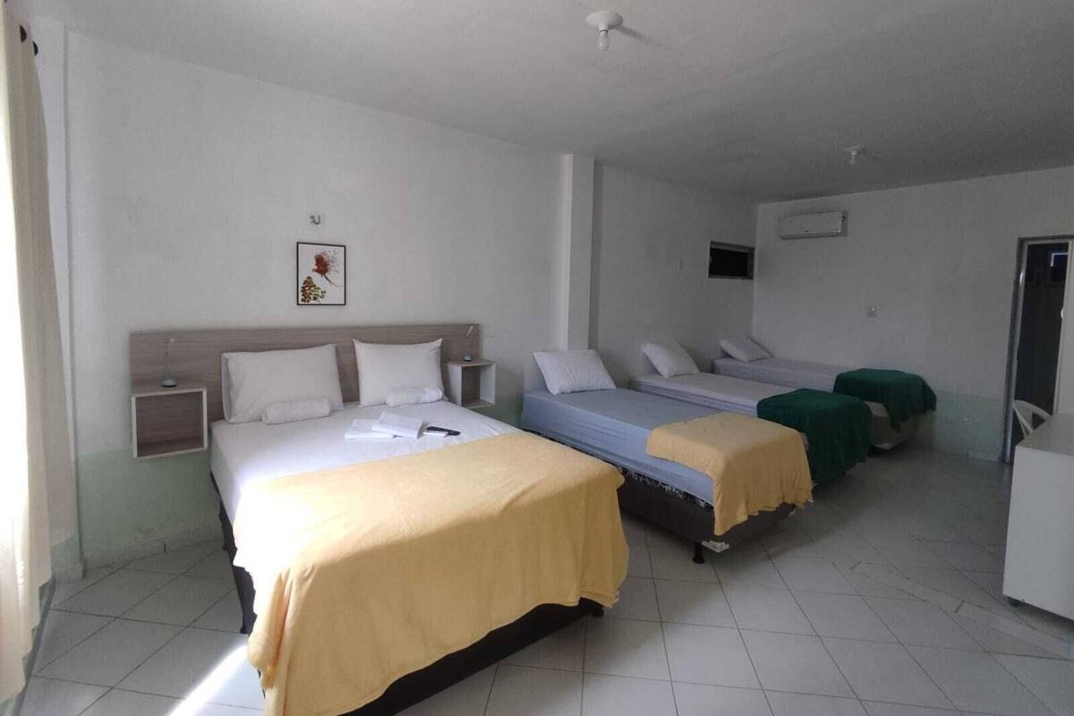 A hotel room with a large bed, two smaller beds, a bedside table, a wall-mounted air conditioner, and a small window.