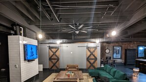 A spacious room with a large flat-screen TV mounted on a white brick wall, a ceiling fan, and a rustic wooden table with chairs. - Industrial urban Loft within 5 minutes from downtown (Highland Park)