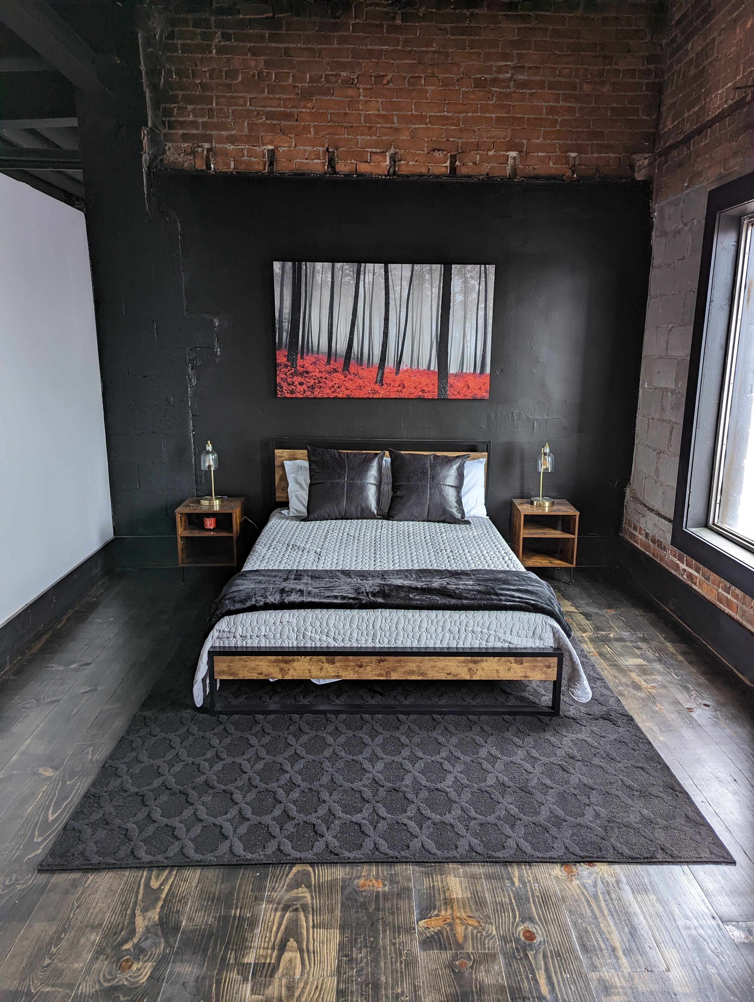 A modern bedroom with a large bed, a black and white forest-themed painting above it, and a brick wall.