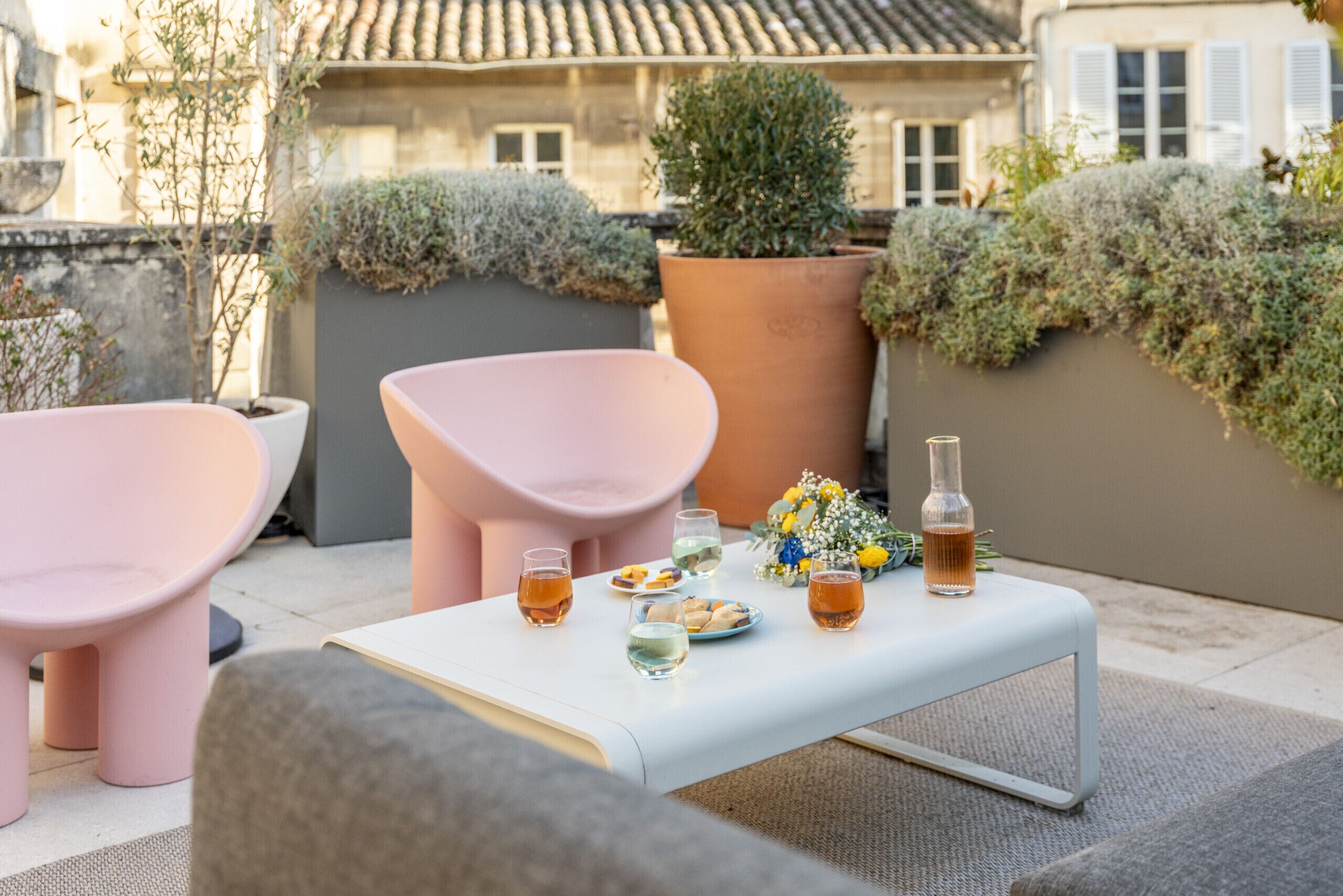 Una terraza con una mesa blanca dispuesta con bebidas, platos y un ramo de flores. Hay dos sillas rosas y un sofá gris.