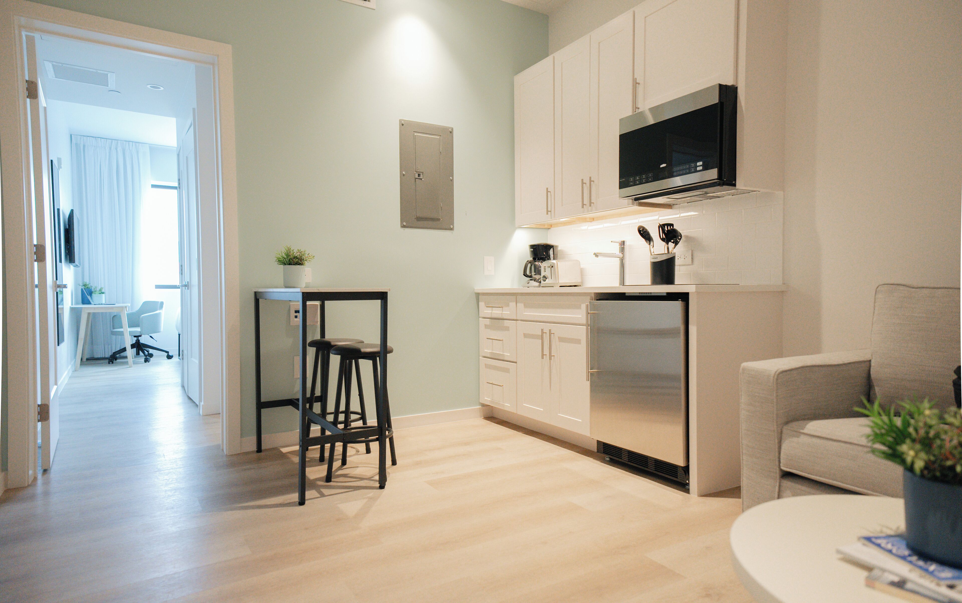 A modern kitchen with white cabinets, a built-in microwave, and a coffee maker. There is a small dining area with a table and chairs.