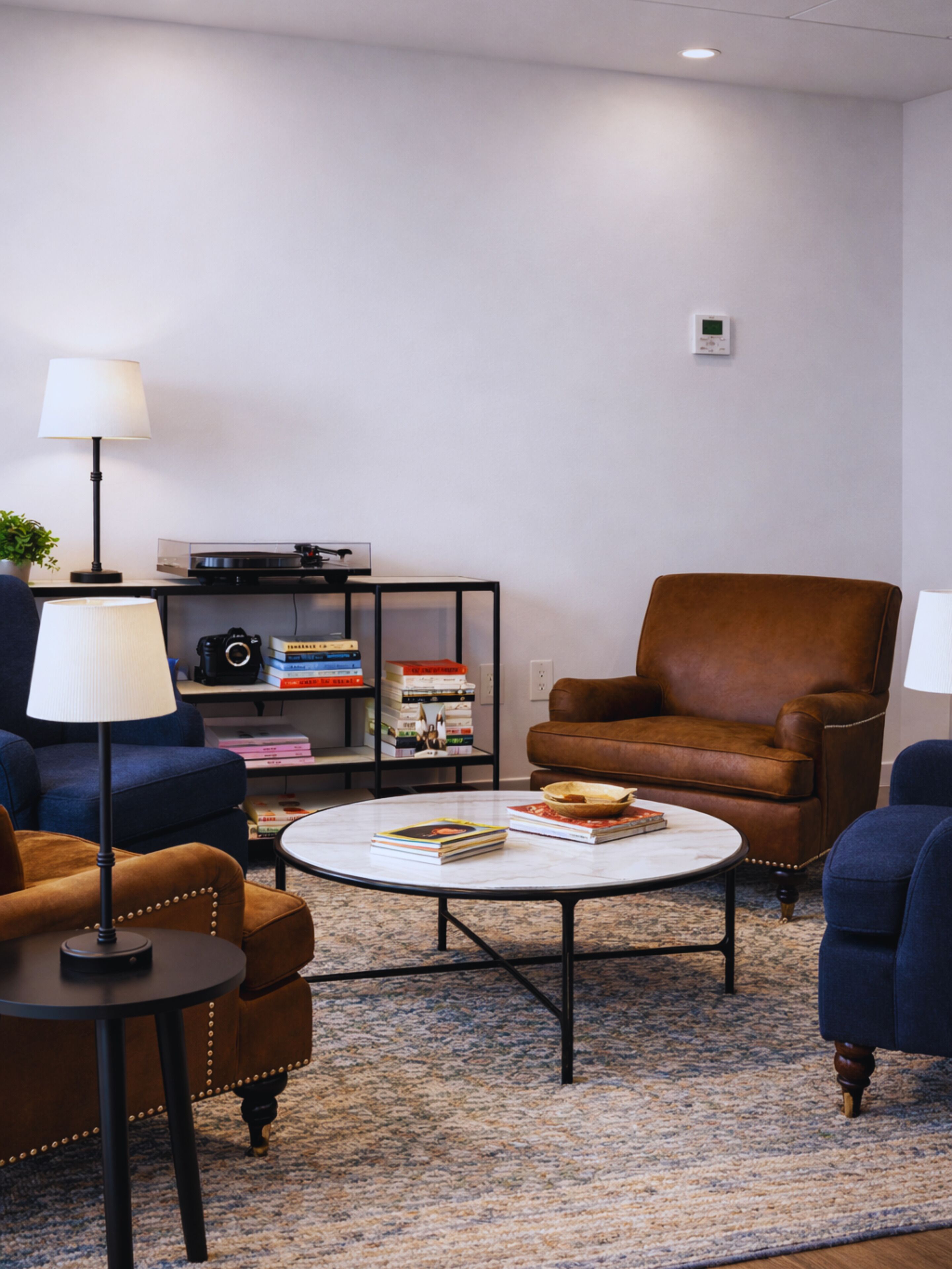 A living room with a brown leather armchair, a blue armchair, a round coffee table, and a bookshelf with books and a camera.
