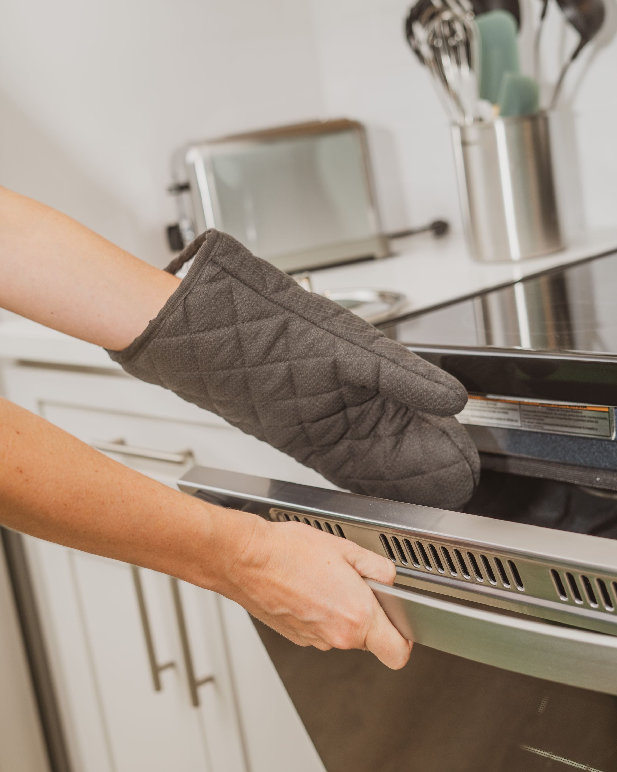 Une personne portant un manique de cuisine matelassé de couleur sombre, qui tend la main vers l’intérieur d’un four en acier inoxydable.