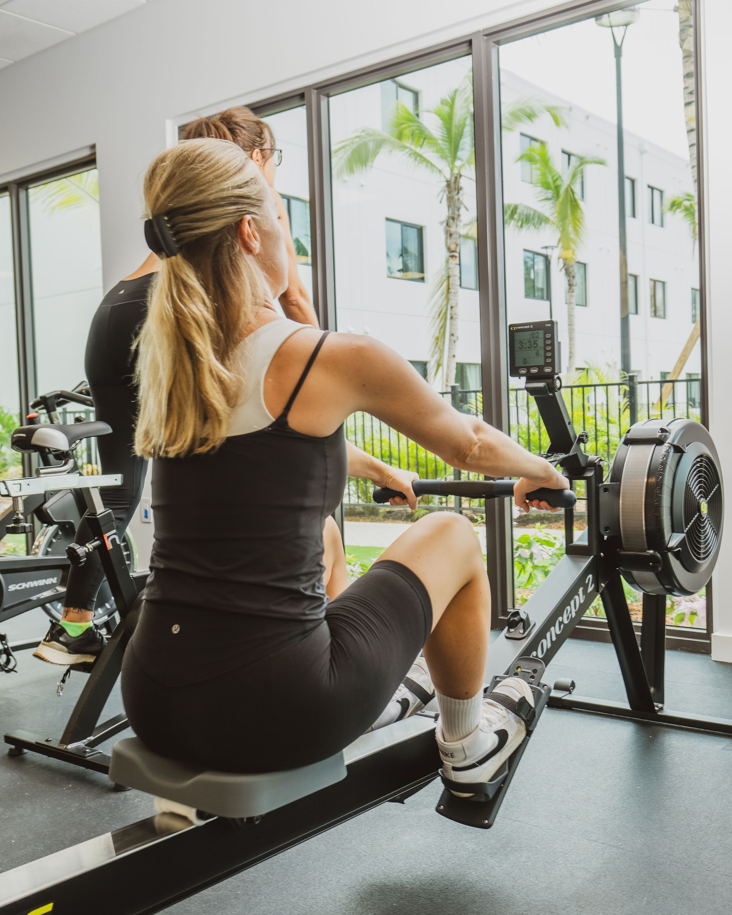Una mujer usando una máquina de remo en un gimnasio con ventanas amplias y vistas a palmeras y edificios.