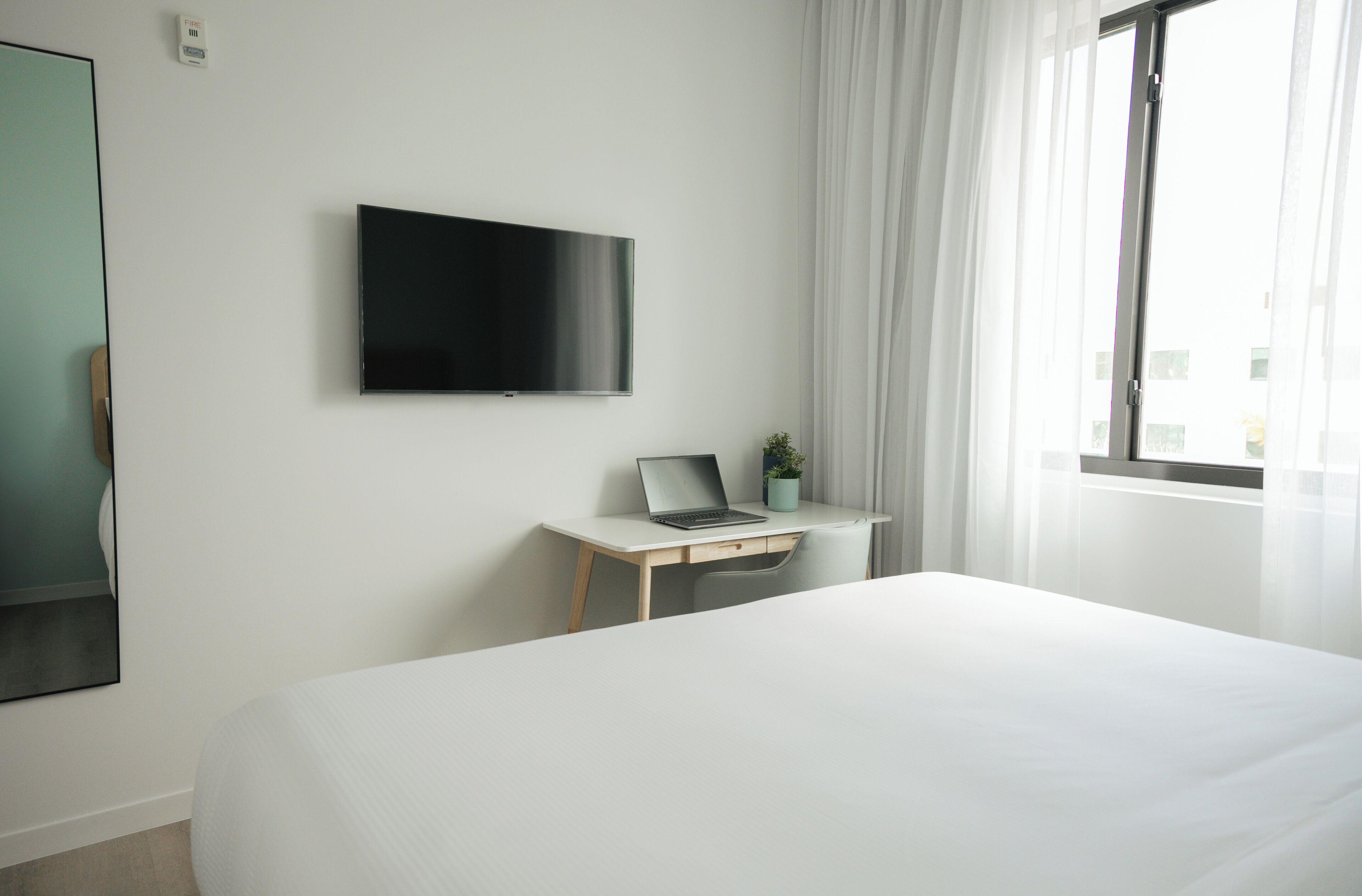 Una habitación de hotel con cama, escritorio con computadora portátil, televisor y ventana con cortinas.