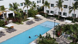 A swimming pool with lounge chairs and umbrellas, surrounded by palm trees and a building with multiple windows.
